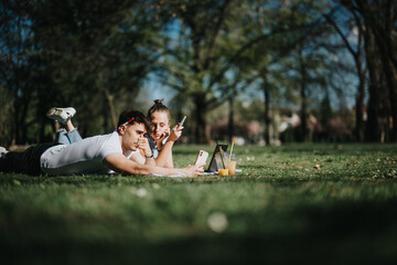 A young couple lies on the grass at the park, closely sharing a joyful moment, taking selfie with...