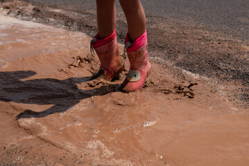 A girl in pink boots is standing in a muddy puddle. The water is brown and murky, and the boots are covered in mud. The scene has a playful and lighthearted mood