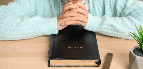 Beautiful young Asian woman with Bible and cross praying at table in living room