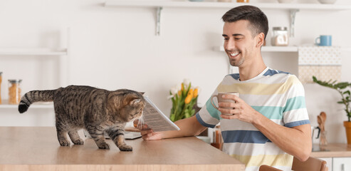 Young man with cute cat reading newspaper in kitchen