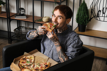 Handsome tattooed young man eating tasty pizza and sitting on armchair at home