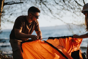 Two friends collaborate to set up an orange tent at a lakeside camping spot, indicating teamwork...