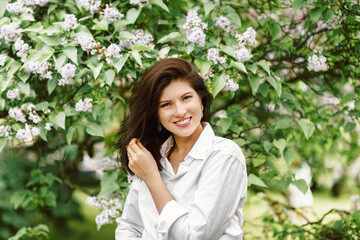 Face and cute smile wearing casual white blouse or shirt in the garden among blooming apple tree branches with small flowers and green leaves looking at camera in sunny day