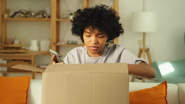 Unhappy african woman unpack delivered box receiving damaged shipping problem in postal service. Annoyed confused customer receiving wrong parcel talks to customers support. Bad delivery services