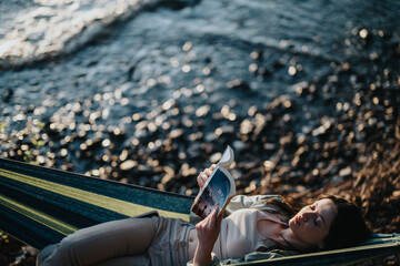 Carefree young girl relaxing with a book in a hammock outdoors, enjoying nature and sunset by a...