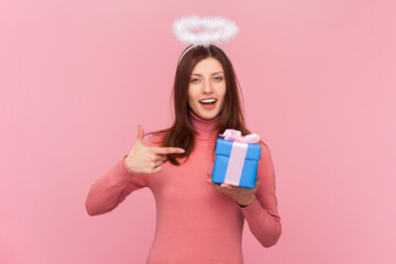 Adorable cheerful woman with brown hair and nimb over head holding and pointing at blue wrapped present box, wearing rose turtleneck. Indoor studio shot isolated on pink background