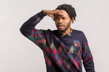 Portrait of serious attentive handsome african-american man with dreadlocks and beard keeps hand near forehead, looking far with interest. Indoor studio shot isolated on gray background.