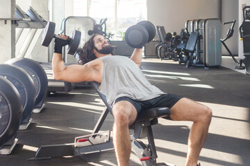 Portrait of strong hard working man with long curly hair working out in gym, doing exercising on bench and holding dumbbells with raised arm, training biceps. Indoor shot.