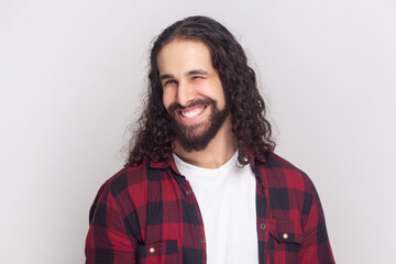 Bearded man with long curly hair in checkered red shirt winking while having good mood smiling,...