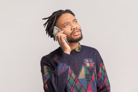 Portrait of funny positive playful african-american man with dreadlocks and beard standing with dollar banknotes near ear, pretending talking on phone. Indoor studio shot isolated on gray background.