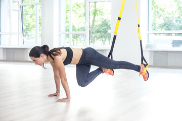 Portrait of athletic strong woman wearing black top and leggings in plank position using fitness straps bending knees while legs hanging. Indoor shot with window on background - obrazy, fototapety, plakaty
