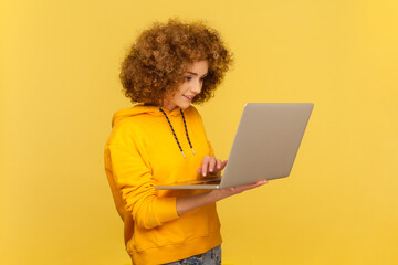 Side view portrait of smiling happy woman with Afro hairstyle holding laptop, typing on keyboard,...