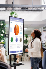 Shopper choosing clothes on display, using touch screen kiosk monitor in trendy mall boutique....