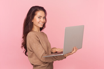Side view portrait of beautiful woman with wavy hair, working on notebook, typing on keyboard, looking smiling at camera, wearing wearing brown pullover. Indoor studio shot isolated on pink background