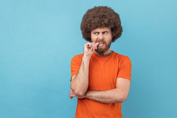 Portrait of nervous terrified man with Afro hairstyle wearing orange T-shirt biting his fingers...