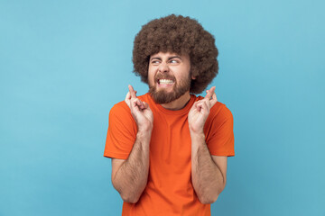 Portrait of man with Afro hairstyle in orange T-shirt standing with crossed fingers for good luck,...