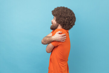 Side view of selfish narcissistic man with Afro hairstyle embracing himself and smiling with expression of great ego, pleasure and self-esteem. Indoor studio shot isolated on blue background. - obrazy, fototapety, plakaty