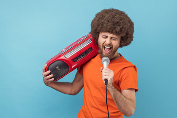 Portrait of excited man with Afro hairstyle wearing orange T-shirt holding tape recorder and...