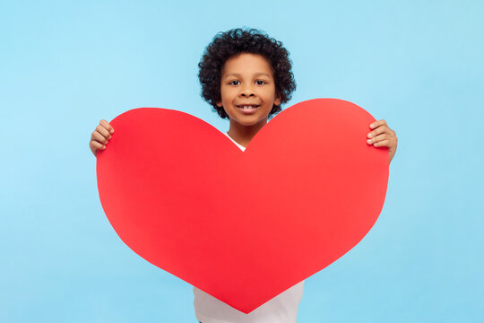 Portrait of charming cute smiling little boy with curly hair holding big red hear demonstrating feelings to his classmate. Indoor studio shot isolated on blue background.