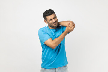 Portrait of upset unhealthy unshaven man wearing blue T- shirt standing touching his injured elbow, frowning face from terrible pain. Indoor studio shot isolated on gray background.