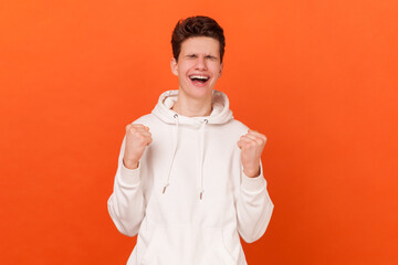 Portrait of happy delighted young man wearing white hoodie clenches fists, feels triumph, rejoices...