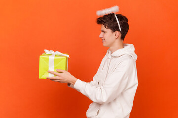 Portrait of smiling happy young man with nimb above his head wearing white hoodie congratulating somebody with birthday, giving present. Indoor studio shot isolated on orange background.