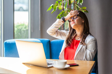 Portrait of unhappy woman working on laptop having job troubles showing facepalm gesture being stressed, wearing jacket and re shirt. Indoor shot, cafe or office background.
