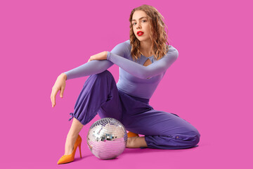 Beautiful young stylish woman with disco ball sitting on purple background