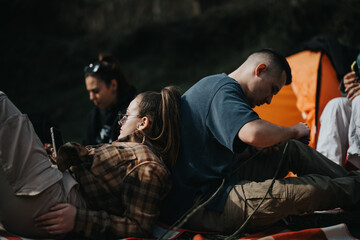 A group of young adults relaxes at a campsite, with one person checking a phone, highlighting a...