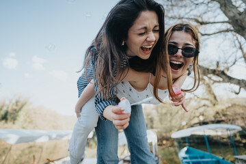 Two young women share a piggyback ride, laughing joyously while blowing bubbles in a sunny outdoor...