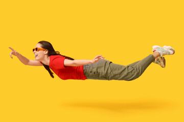 Joyful young woman in sunglasses flying and pointing at something on yellow background