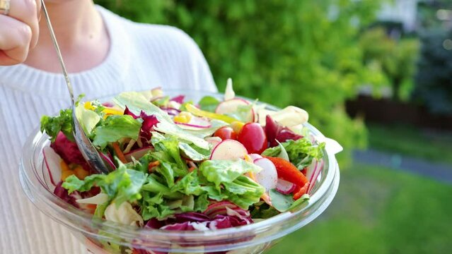 Woman eating, cooking vegetable, vegetarian salad outdoors in summer. Mixing a fresh vegetables vegan salad. Bowl of romaine lettuce, arugula, tomato, paprika, red onion, dressing with olive oil