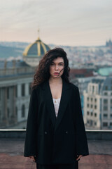 Attractive young curly brunette woman in a black jacket and black pants stands on a rooftop against a historic building during a magical sunset.