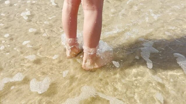 Child girls legs walking barefoot on beach coastline in ocean shallow water. Atlantic ocean beach and child walking in sea or ocean waves. 4K resolution video
