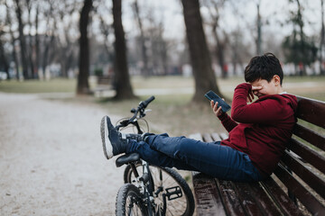 A joyful young boy takes an active rest on a park bench, casually lying with his bike nearby and...