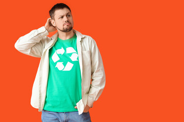 Confused young man in t-shirt with recycling logo scratching head on orange background. Ecology...