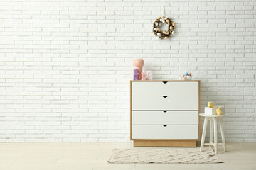 Beautiful Easter wreath decorated with eggs on white brick wall near chest of drawers in room