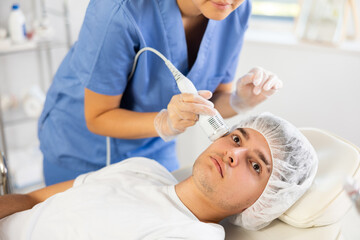 Young woman cosmetologist performs facial whitening procedure with apparatus young male patient