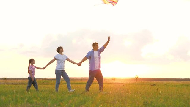 Mother father and daughter playing flying kite at sunset natural field green grass. Happy family parents and girl kid child holding hands relaxing together outdoor meadow flight toy entertainment