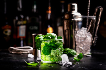 Green summer alcoholic cocktail drink with dry gin, sugar syrup, lemon, green basil and ice, dark bar counter background - 792153313