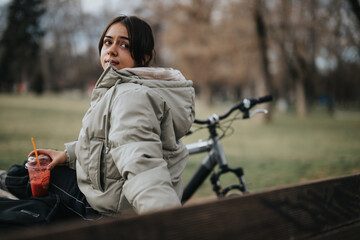 A carefree young girl sits with her bicycle in the park, savoring a refreshing drink during a...