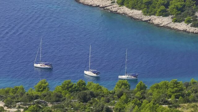 AERIAL: Three luxury sailboats are anchored next to a rugged Mediterranean coast, symbolizing the epitome of carefree summertime vacation. Sailboats moored on the shores of scenic Hvar island, Croatia