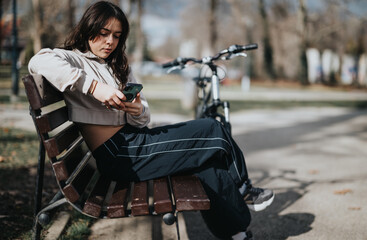 Fototapeta na wymiar Focused young adult browsing her phone while sitting on a wooden bench in a sunny park, with a bicycle leaned nearby.