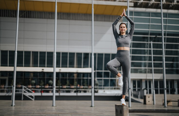 An athletic woman performs a yoga pose outdoors in front of a modern building, embodying...