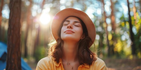 Young woman standing among trees in the woods, wearing a hat with her eyes closed, taking deep breaths of fresh air.