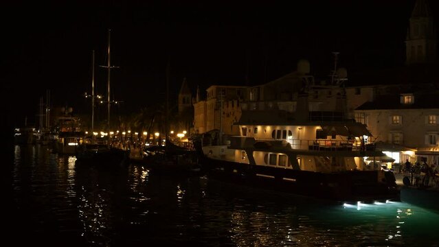 An evening view of moored sea vessels along the bustling city harbour promenade filled with a strolling crowd of people. Yellow street lights reflect in calm harbor water of charming seaside town.