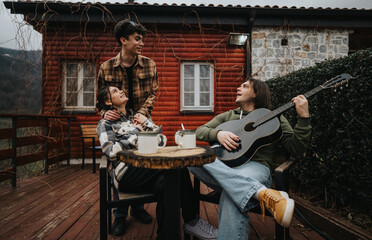 Three young adults share a moment of happiness with a guitar on a wooden cabin porch, exuding warmth and togetherness.