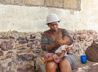 village african woman feeding her baby outdoors, sited in front of the house in the yard