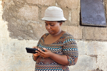 young african chubby woman with a hat holding her cellphone in the poor township, informal settlement