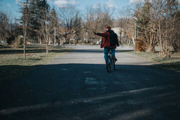 A person on a bicycle wearing a red jacket and beanie with a backpack pointing to the side in a serene park setting.